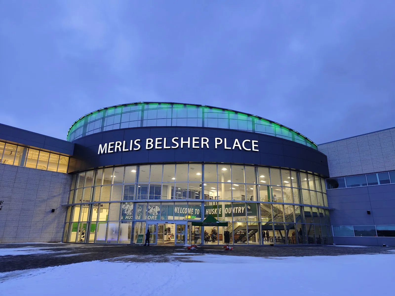The curved glass entrance with MERLIS BELSHER PLACE in white letters and a green-lit upper band, at dusk with snow in front. — Merlis Belsher Place