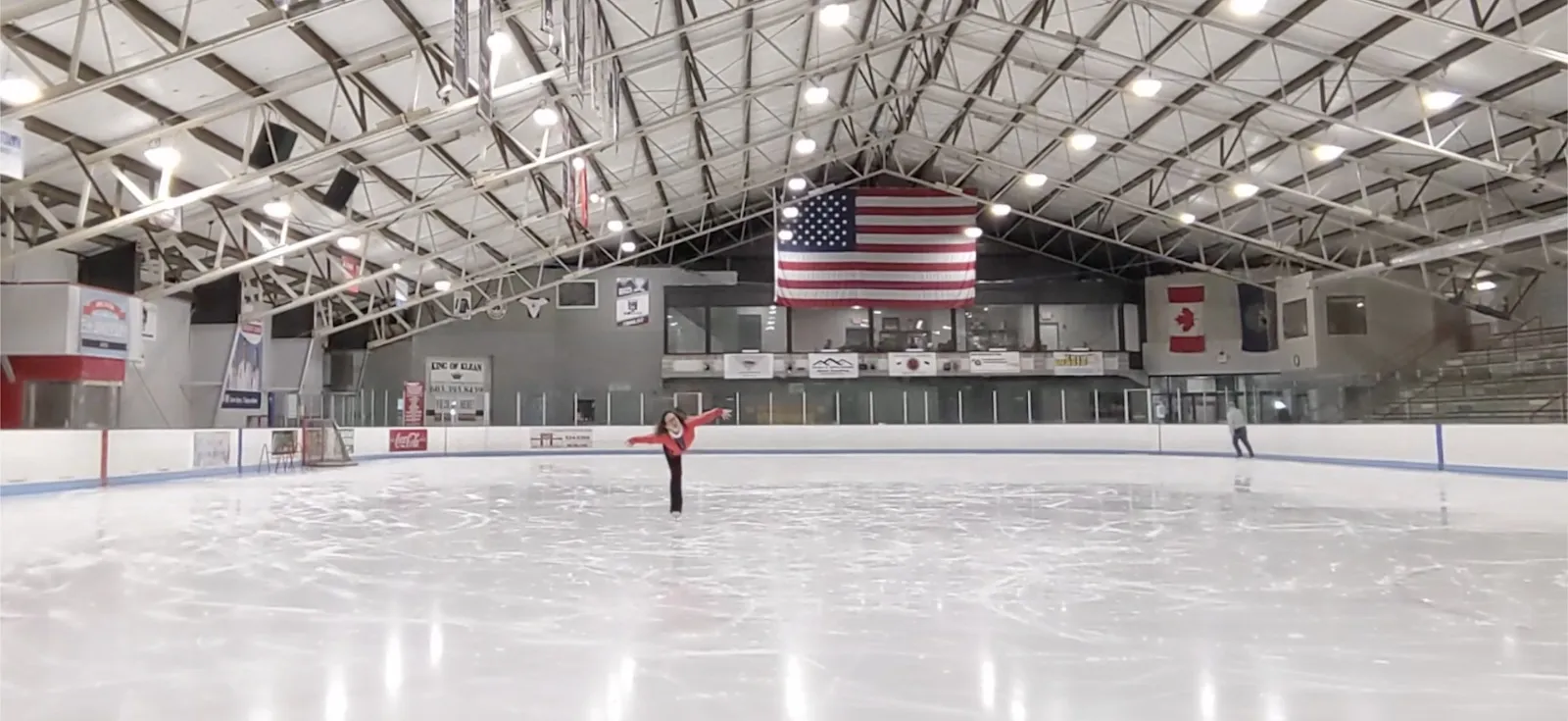 Una vista amplia de la pista con una bandera de Estados Unidos en la pared del fondo, un solo patinador cerca de las vallas bajo un techo de armaduras de acero con banderines. — Merrill Fay Arena