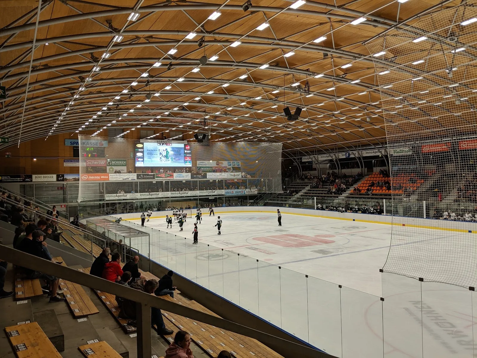 Die Eisfläche des Messestadions Dornbirn über Glas und Netz von den Rängen aus gesehen, mit einigen Schlittschuhläufern auf dem Eis und dem geschwungenen Holzdach darüber. – Messestadion Dornbirn