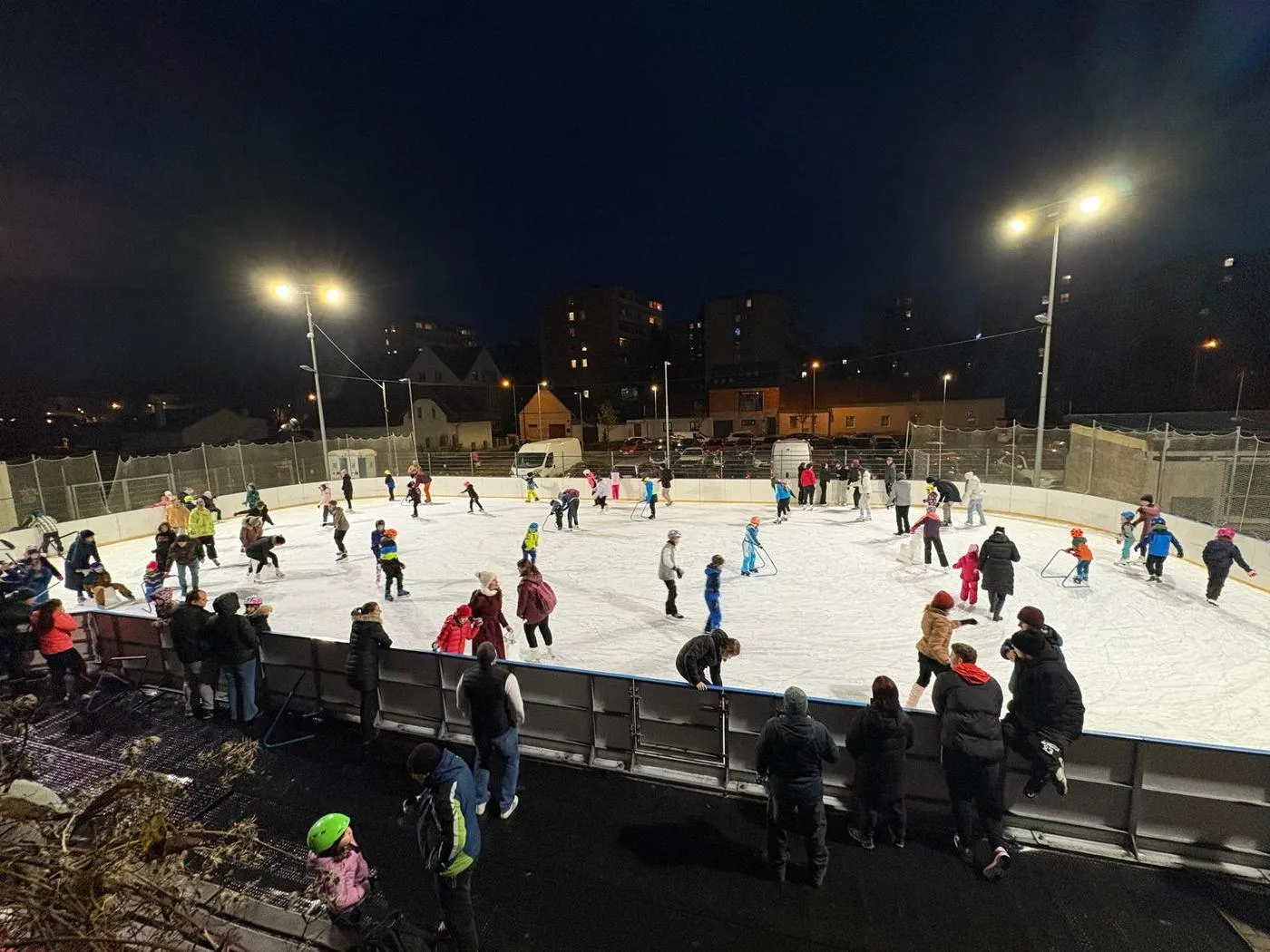 The floodlit rink in Brandýs nad Labem-Stará Boleslav at night, with families skating inside a fenced pitch and houses on the hill beyond. — Městské kluziště Brandýs nad Labem-Stará Boleslav
