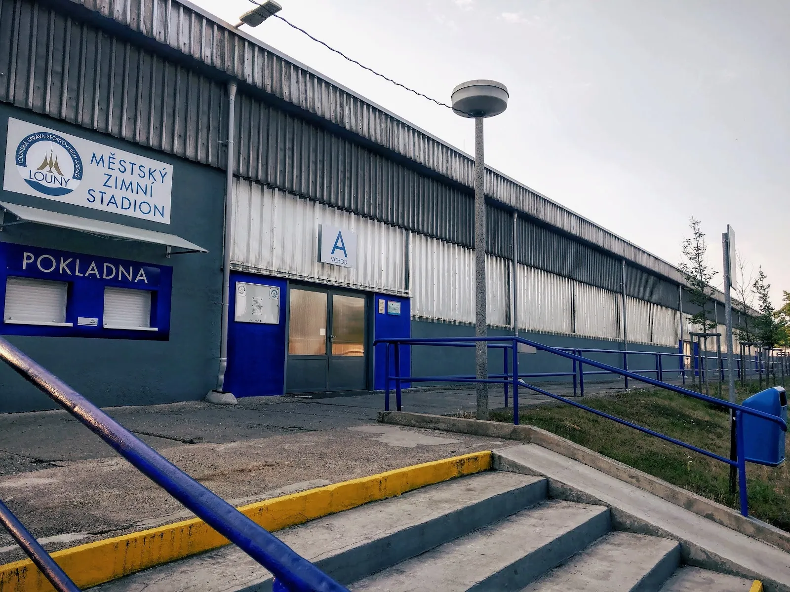 Vchod s pokladnou Městského zimního stadionu v Lounech s nápisem stadionu a okénkem POKLADNA na dlouhé modrošedé stěně haly – Městský zimní stadion Louny