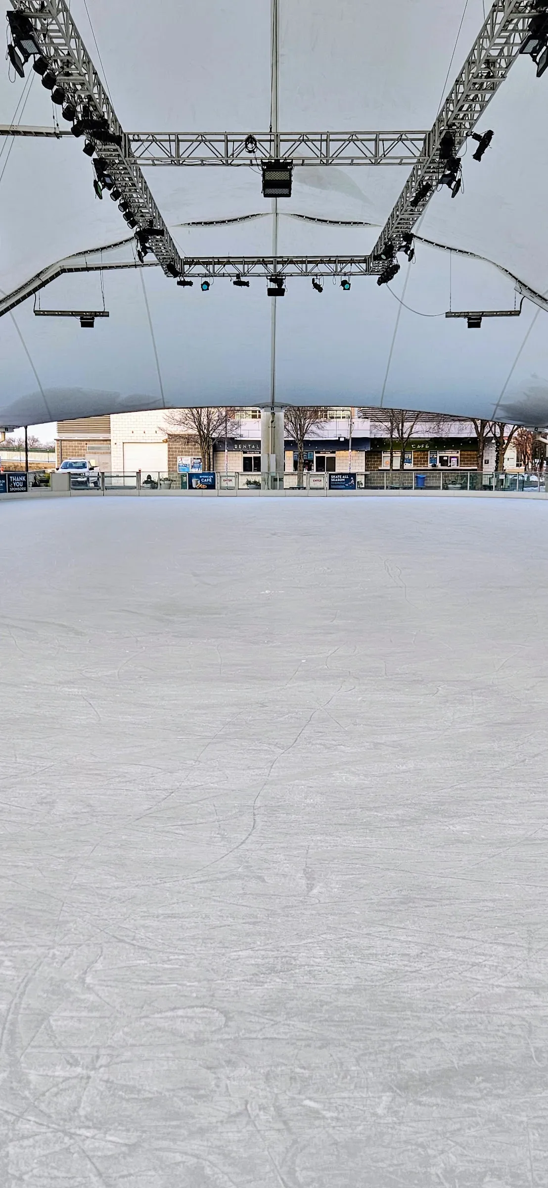 The empty ice of the MetroParks Ice Rink at RiverScape under the white fabric canopy and its lighting truss, with the Dayton riverfront beyond — MetroParks Ice Rink at RiverScape