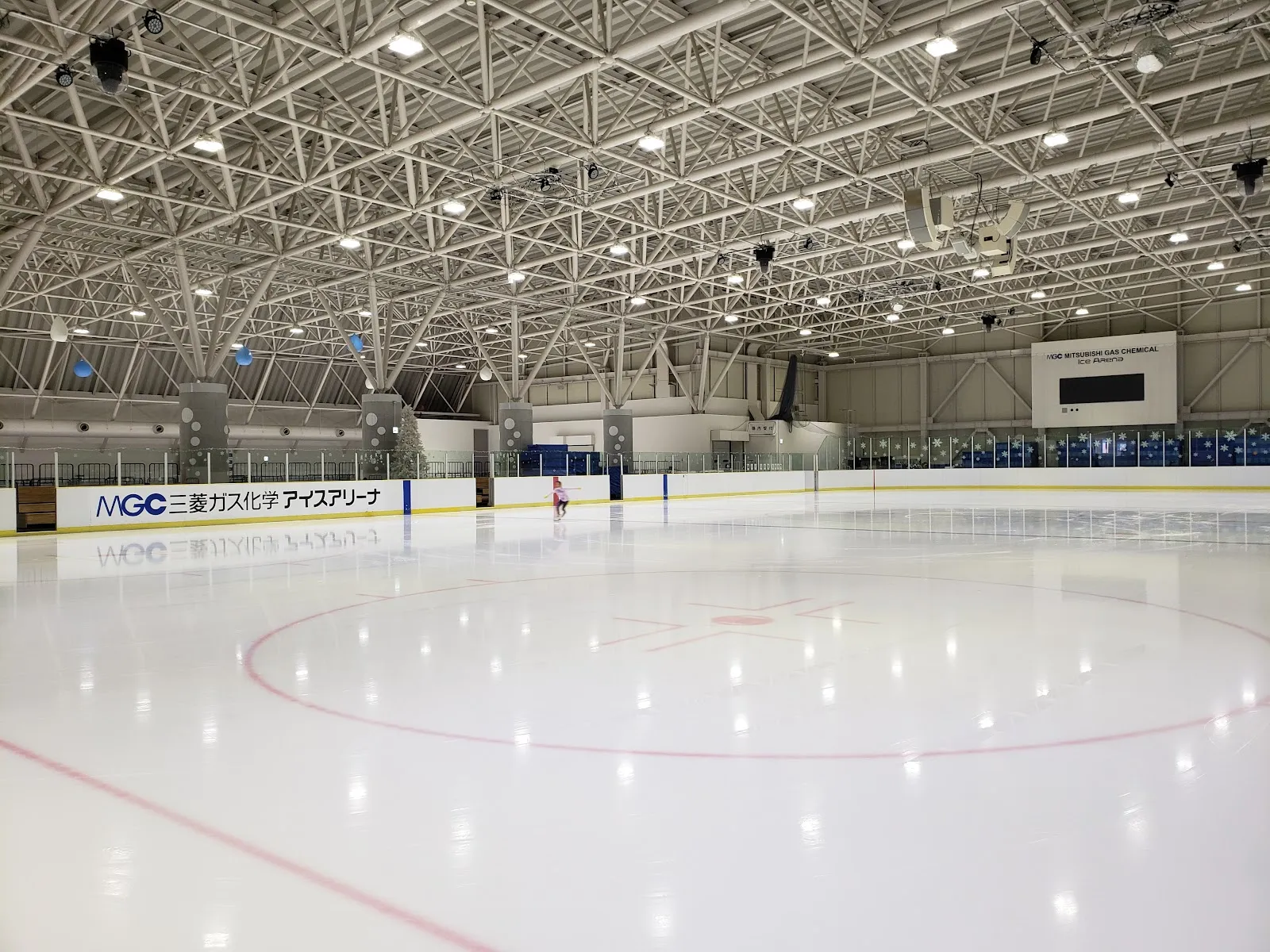 MGC Mitsubishi Gas Chemical Ice Arenaの広い氷面。フェンスにアリーナの名前があり、赤いラインとセンターサークル、白い鉄骨の屋根が見え、氷の向こうに小さな滑走者が1人います — MGC Mitsubishi Gas Chemical Ice Arena