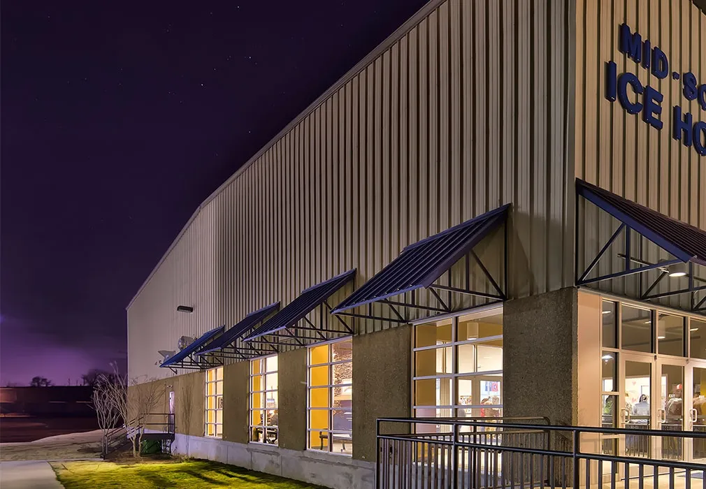 Night view of Mid-South Ice House: a tan metal-sided building with dark blue awnings over lit windows, glass entrance doors and blue 'MID-SOUTH ICE HOUSE' lettering (partly cropped) on the facade. — Mid-South Ice House