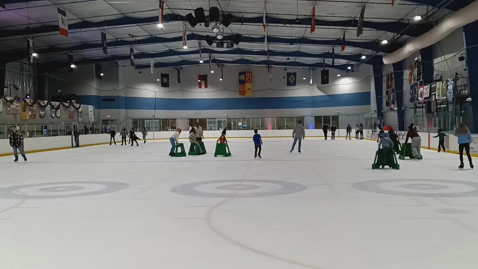 A public session on a wide sheet, skaters spread across white ice with faceoff circles beneath a high roof hung with flags and club banners. — Mid-South Ice House