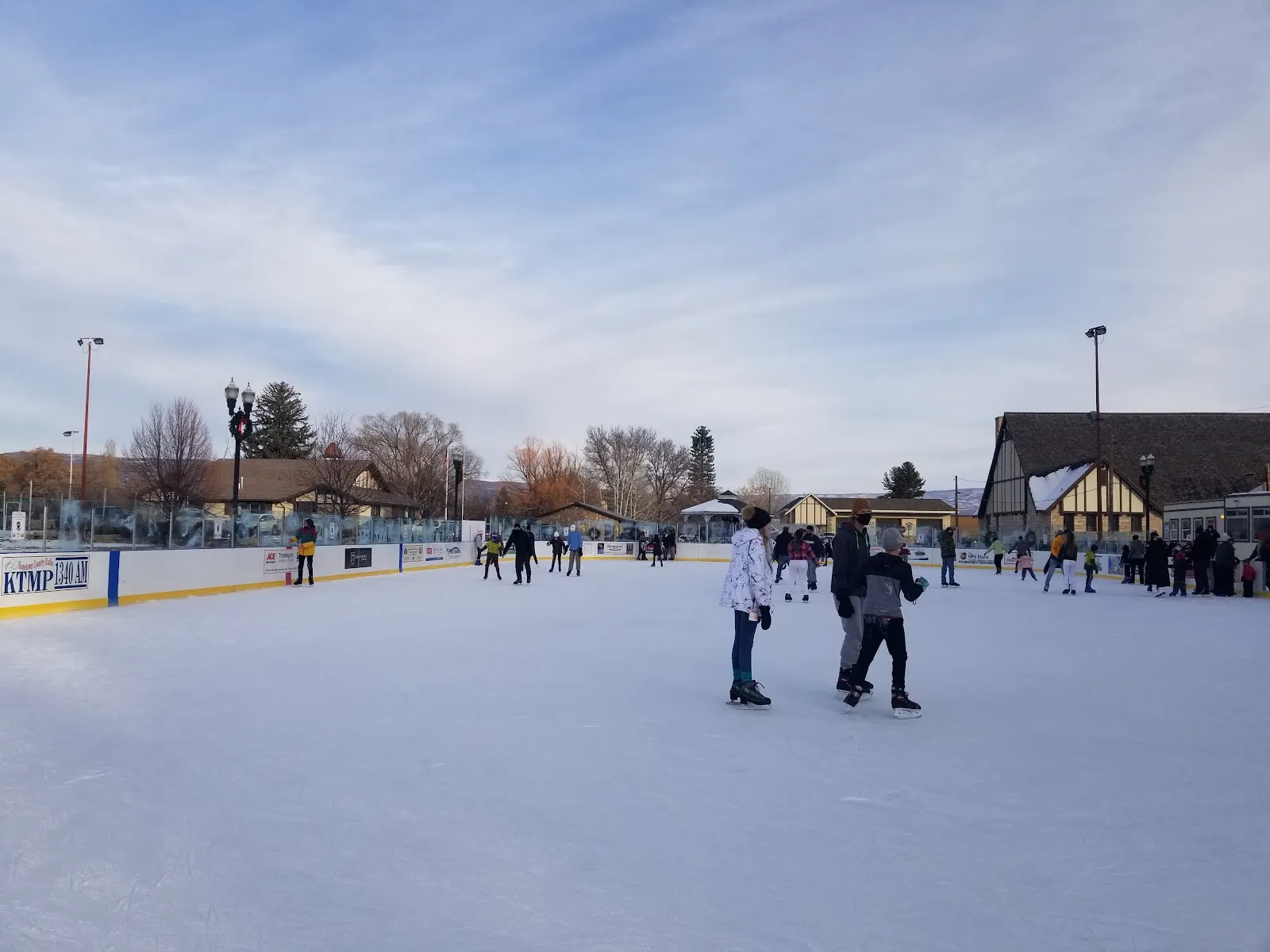 The Midway Ice Rink on the Town Square at dusk, with skaters on the ice, the dasher boards and the Town Square buildings and trees behind. — Midway Ice Rink