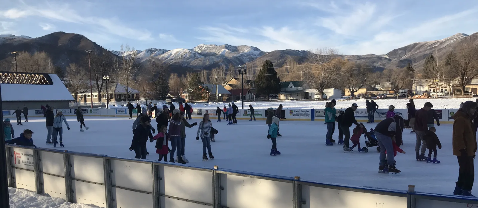 Skaters at a distance on the outdoor ice of the Midway Ice Rink, with the dasher boards in front and the Wasatch mountains behind the Town Square. — Midway Ice Rink
