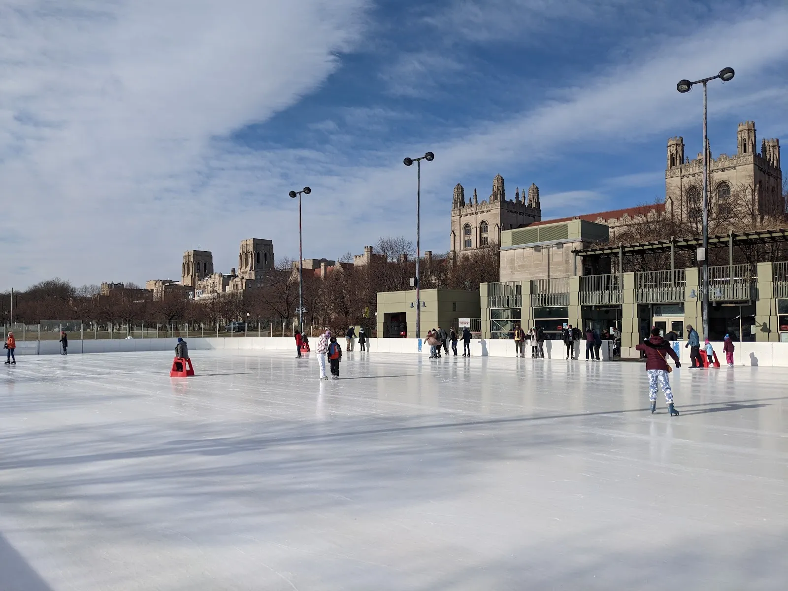La pista y su caseta de calentamiento con fachada de vidrio en el Midway, con torres universitarias góticas detrás. — Midway Plaisance Ice Rink
