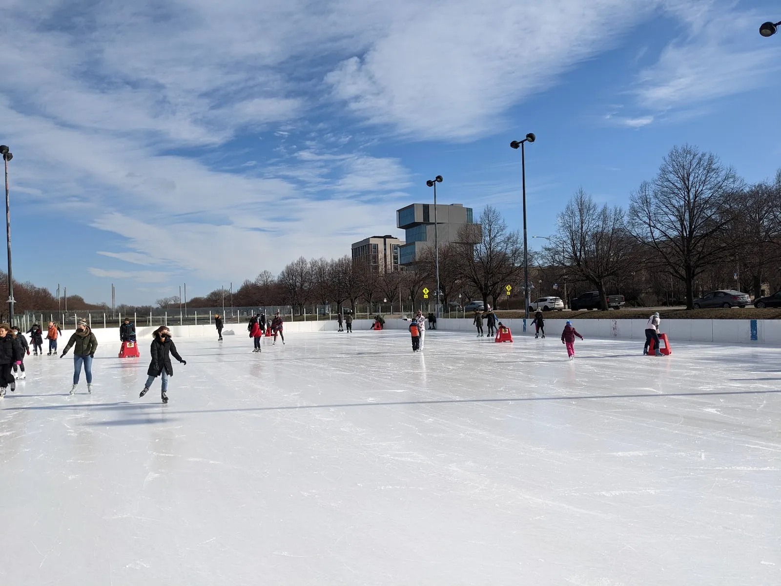 Patinadores repartidos por la pista al aire libre en un soleado día de invierno, con postes de luz, árboles sin hojas y edificios del campus más allá de las vallas. — Midway Plaisance Ice Rink