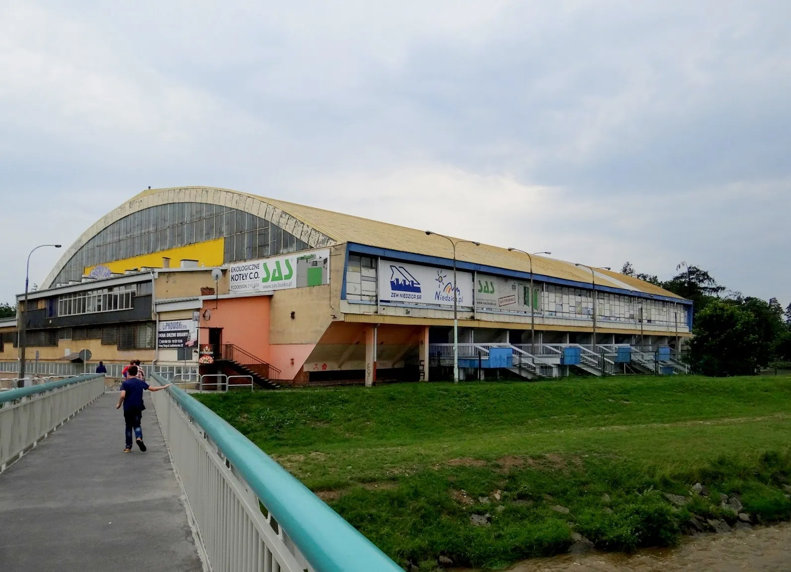 Długa hala lodowa w Nowym Targu z łukowym dachem, widziana z kładki nad trawnikiem, z bladożółtym frontem i szyldami sponsorów. — Miejska Hala Lodowa Nowy Targ