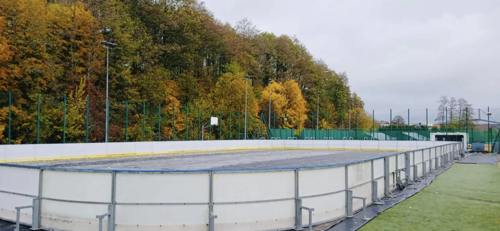 The boarded municipal rink at Sokółka between seasons, seen along the white boards with a football pitch on the right and an autumn-coloured wooded hillside behind. — Miejskie Lodowisko OSiR Sokółka
