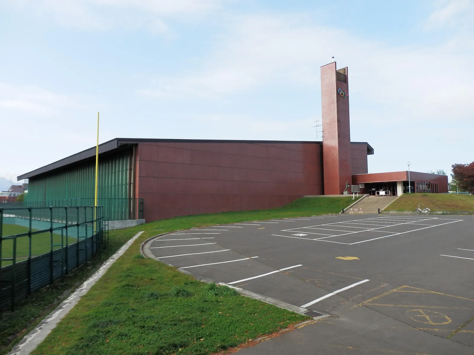 赤茶色の壁の札幌・美香保体育館。入口の上にオリンピックの五輪を掲げた高い塔が立ち、手前には駐車場が広がっている。 — Mikaho Gymnasium Skating Rink