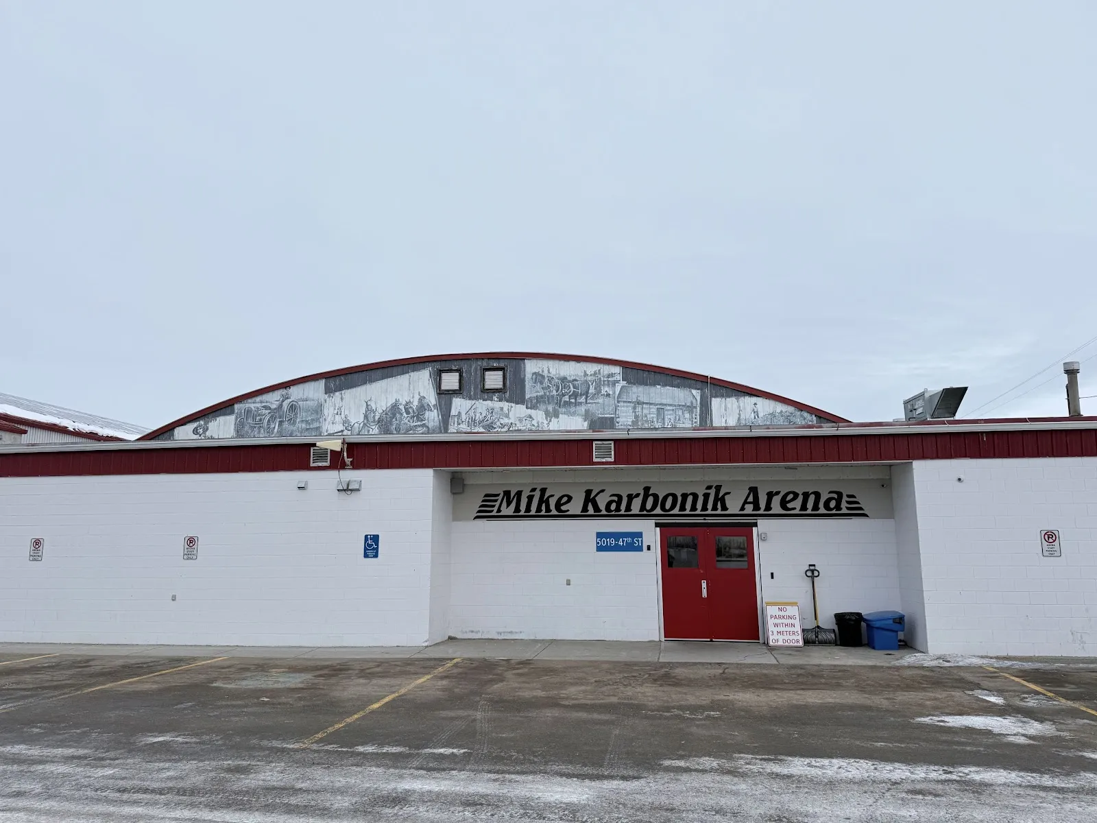 The white arena front with the Mike Karbonik Arena sign over red doors and a mural along the curved roofline. — Mike Karbonik Arena