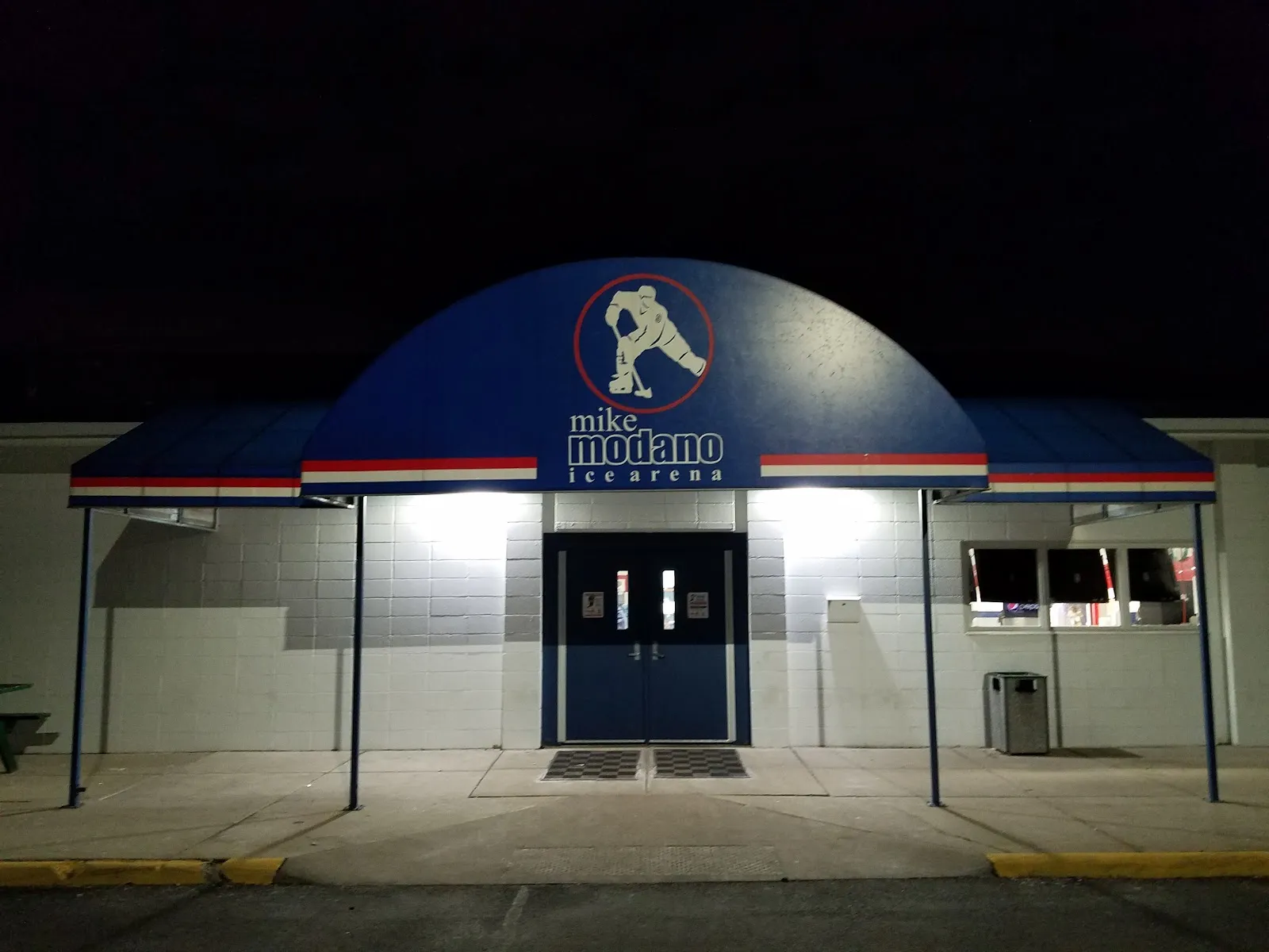 The arena entrance at night: blue double doors under a blue arched canopy with the Mike Modano Ice Arena logo, set in a white block wall. — Mike Modano Ice Arena