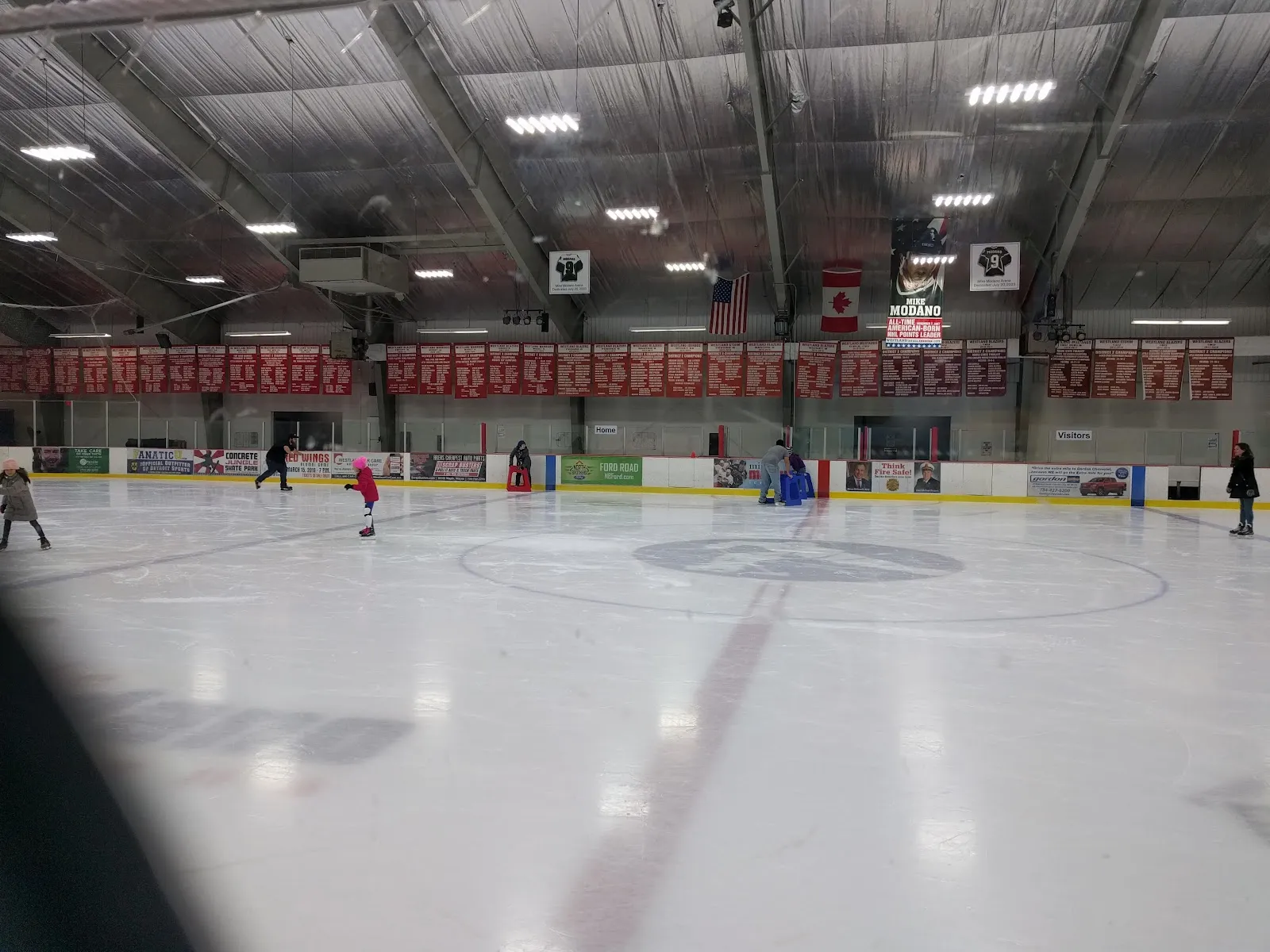 The sheet from behind the glass during an open skate, with a few distant skaters, the centre-ice logo and rows of red and white banners along the far wall. — Mike Modano Ice Arena