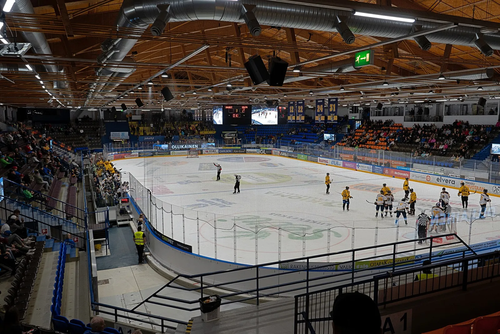 A league game in progress on the ice of Mikkelin jäähalli, seen from the stands, with spectators in the stands and the timber roof overhead. — Mikkelin jäähalli