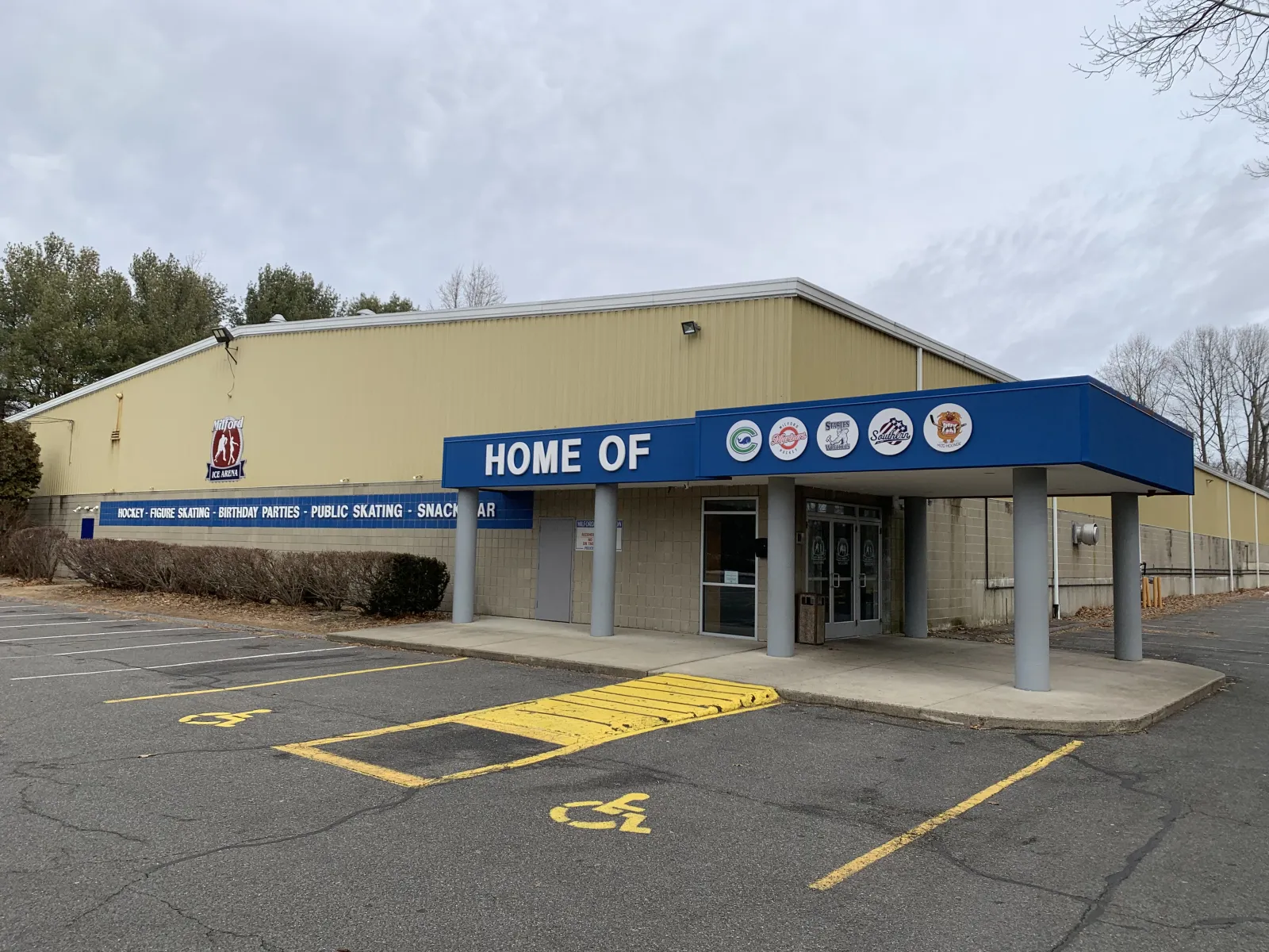 Front entrance of the tan metal building with a blue canopy reading "HOME OF" over team logos, a wall sign reading "HOCKEY - FIGURE SKATING - BIRTHDAY PARTIES - PUBLIC SKATING - SNACK BAR", and the Milford Ice Arena crest on the wall, seen from the parking lot. — Milford Ice Arena