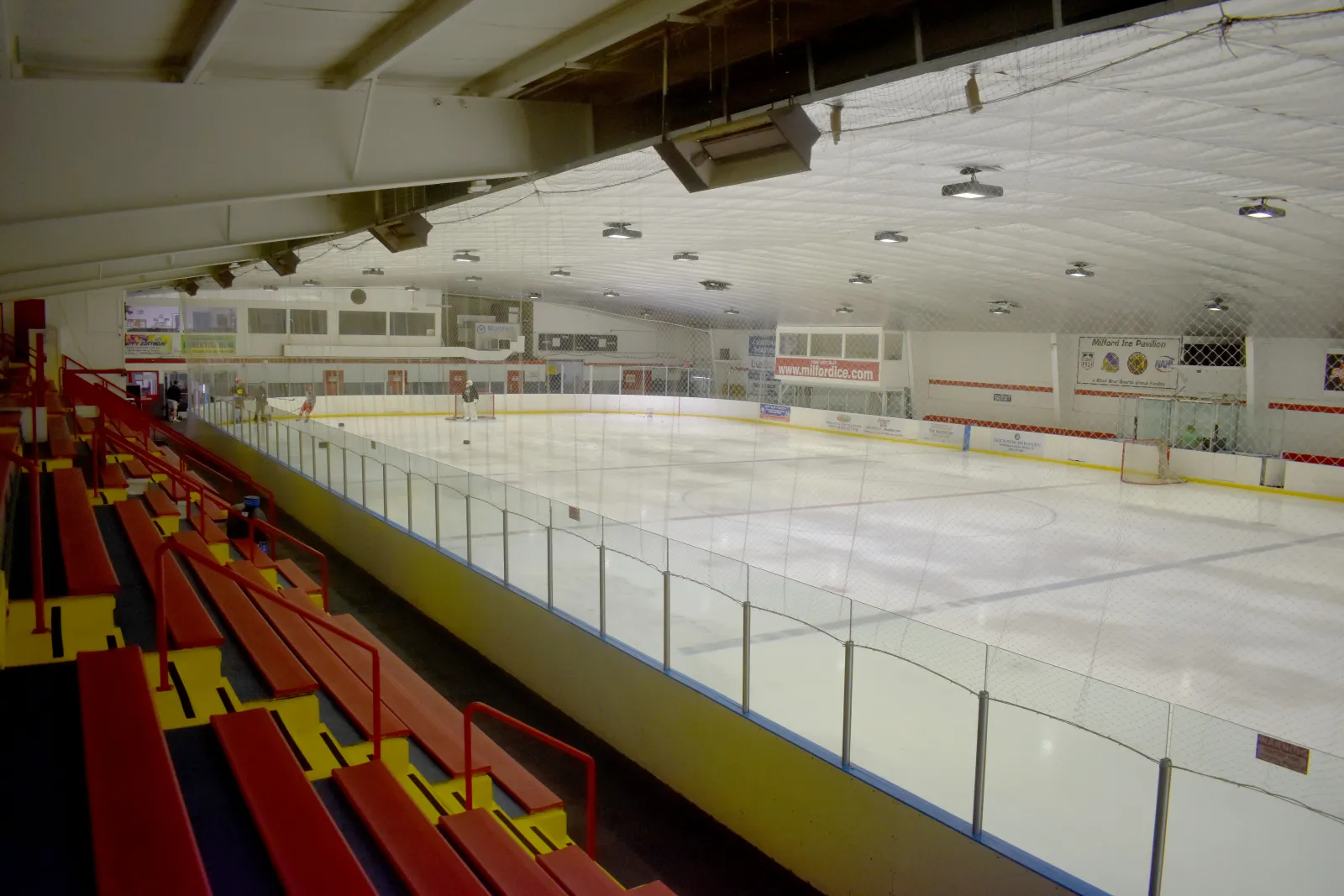 The full ice sheet seen from the red bleachers, with a hockey net, "www.milfordice.com" on the boards and a Milford Ice Pavilion sign beyond. — Milford Ice Arena
