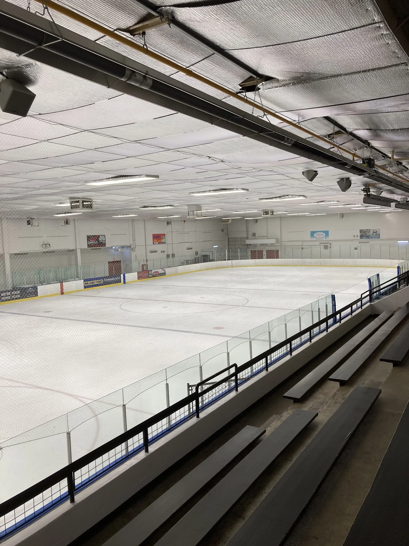 A wide sheet seen from the upper rail, an empty sheet with a pale roof and a long bench along the near side. — Mill Woods Twin Arena