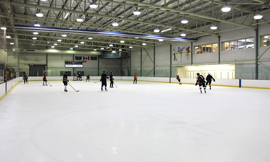 La pista de la arena de Millennium Place con patinadores y jugadores repartidos por el hielo, un borde de vallas con línea amarilla y un alto techo con estructura de acero. — Millennium Place
