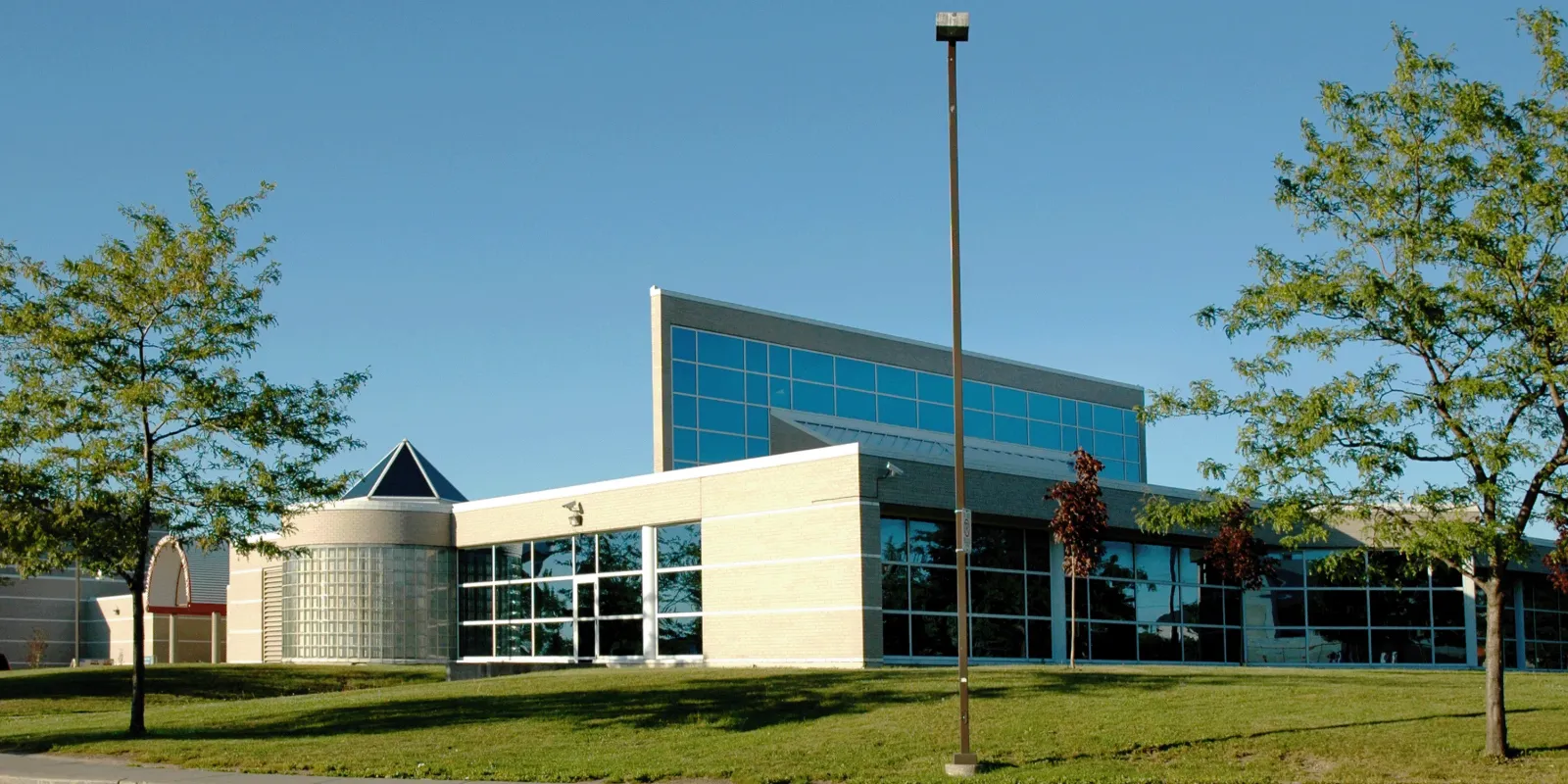 Front entrance of Milliken Mills Community Centre, a beige-brick building with a glass entrance and a taller blue-glass tower section, seen from the parking lot on a clear day. — Milliken Mills Community Centre