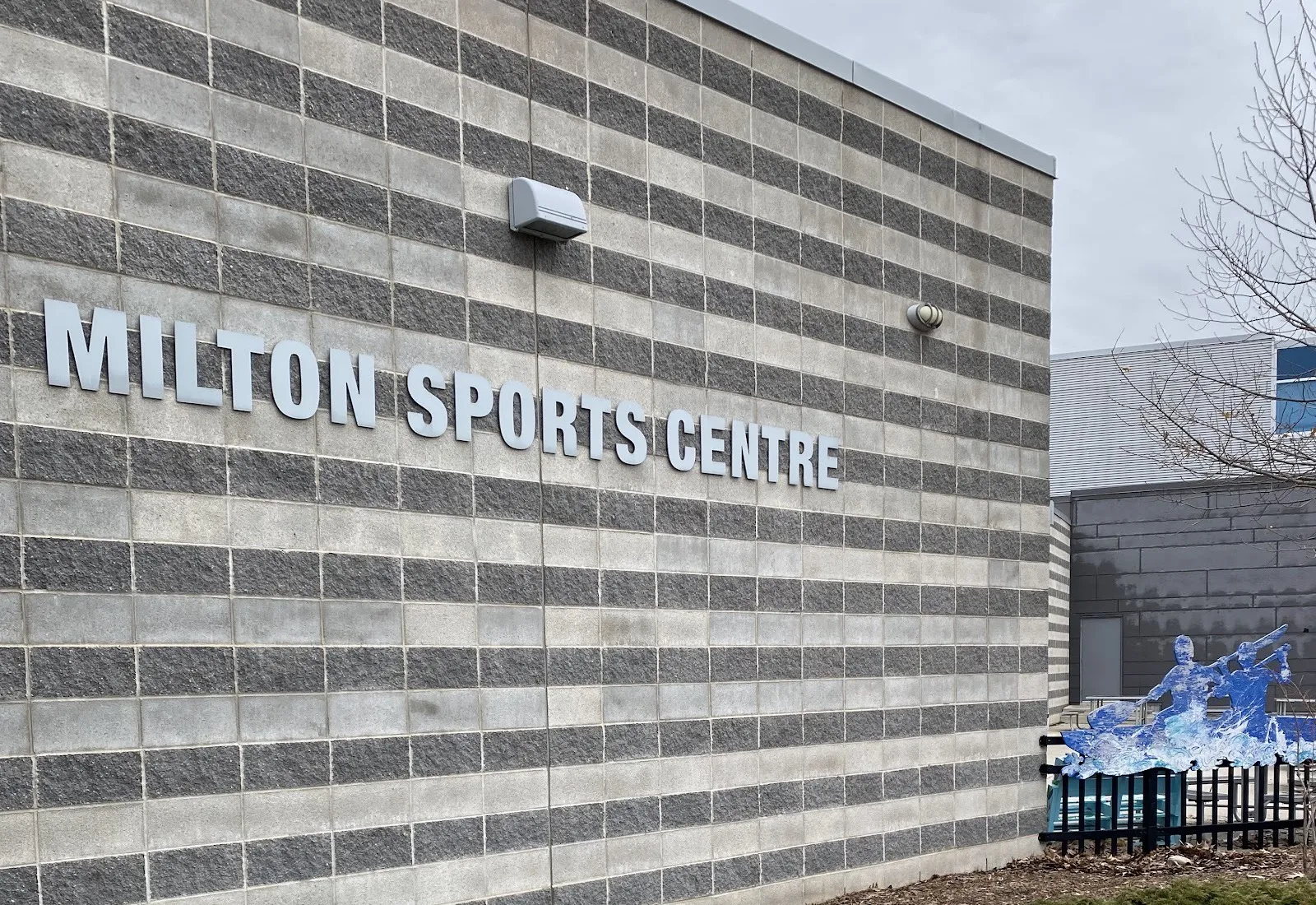 Un muro del Milton Sports Centre de bloques grises en franjas, con el nombre del centro en letras metálicas y el ala más nueva detrás — Milton Sports Centre
