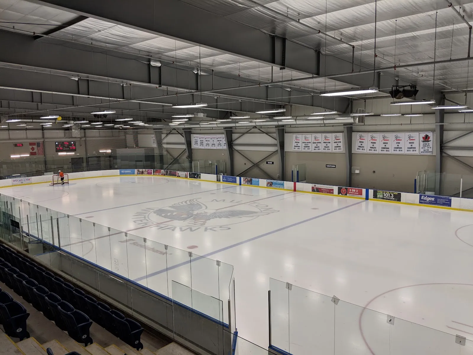 Vista elevada desde las filas superiores de asientos sobre una pista vacía con el logotipo de los Milton Winterhawks en el centro de la pista, cristal a lo largo de las vallas cercanas, mantas de campeonato de la OMHA en la pared del fondo y un patinador colocando una portería en el extremo opuesto. — Milton Sports Centre
