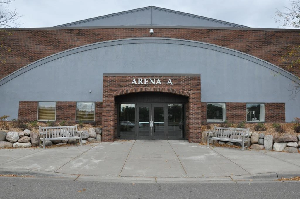 Minnetonka Ice Arena’s Arena A entrance, a brick archway set into the curved grey facade, with benches on either side. — Minnetonka Ice Arena