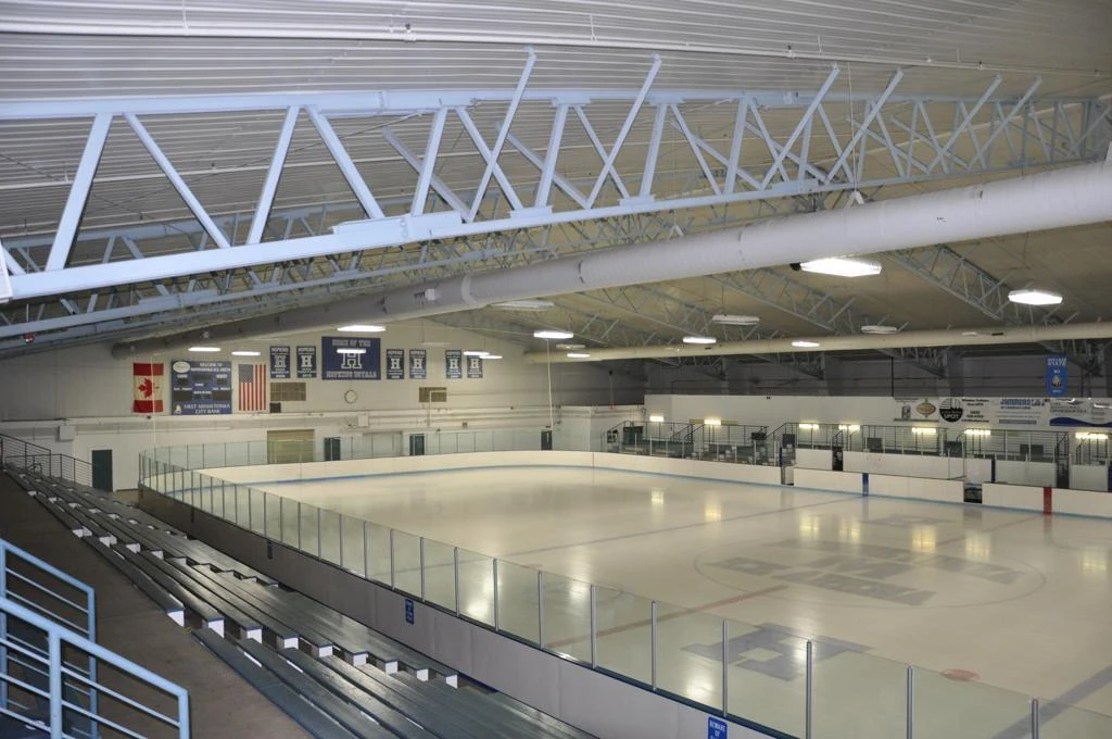 Arena A’s empty ice surface seen from the top of the bleachers, beneath white roof trusses and wall banners. — Minnetonka Ice Arena