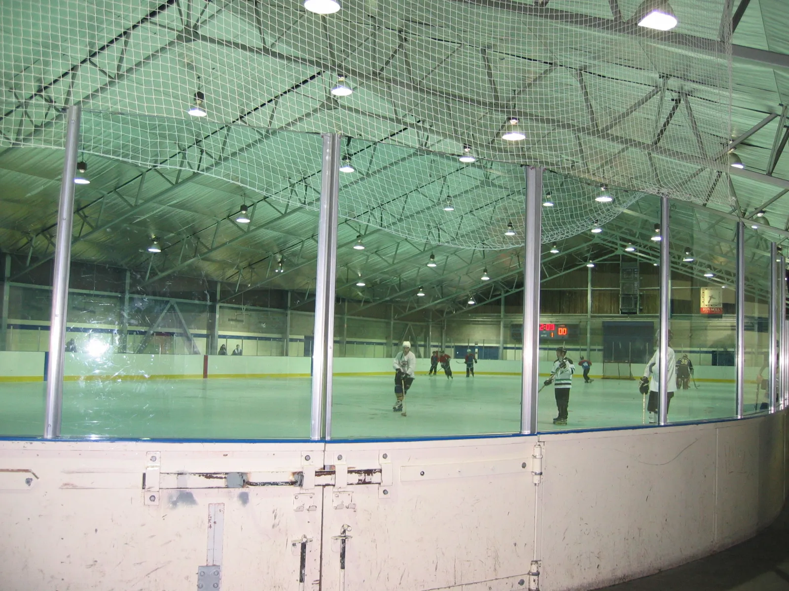 Interior view through the glass at Minoru Arenas, showing hockey players on the ice sheet, the arched steel-truss roof and overhead protective netting. — Minoru Arenas