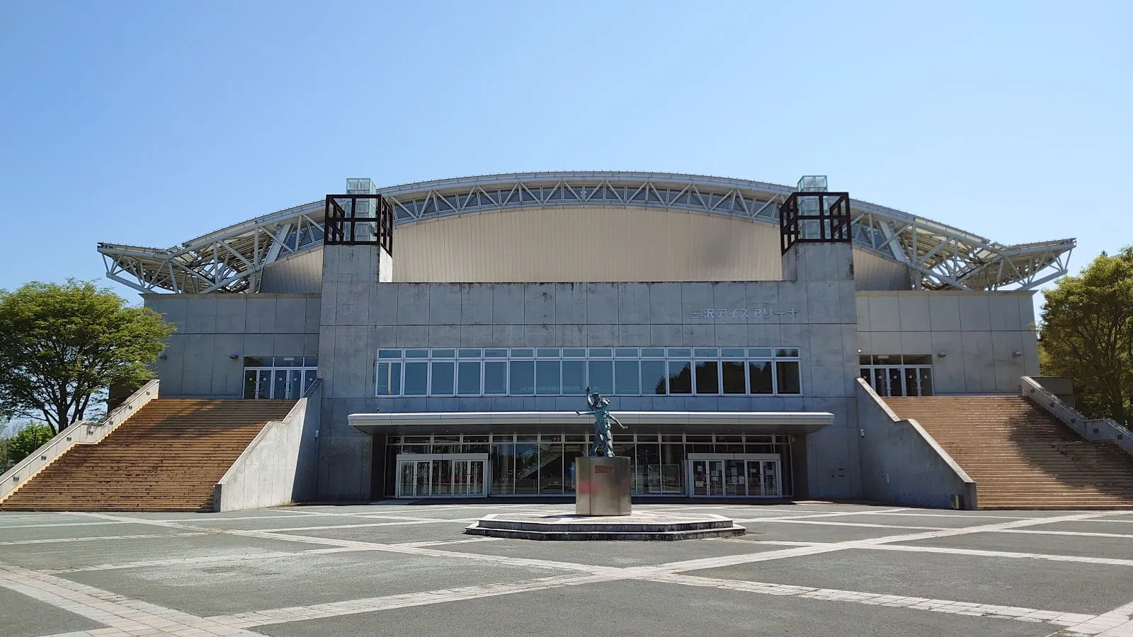 The front of Misawa Ice Arena under its arched steel roof, with wide stone steps either side of a plaza in front of the glass entrance — Misawa Ice Arena
