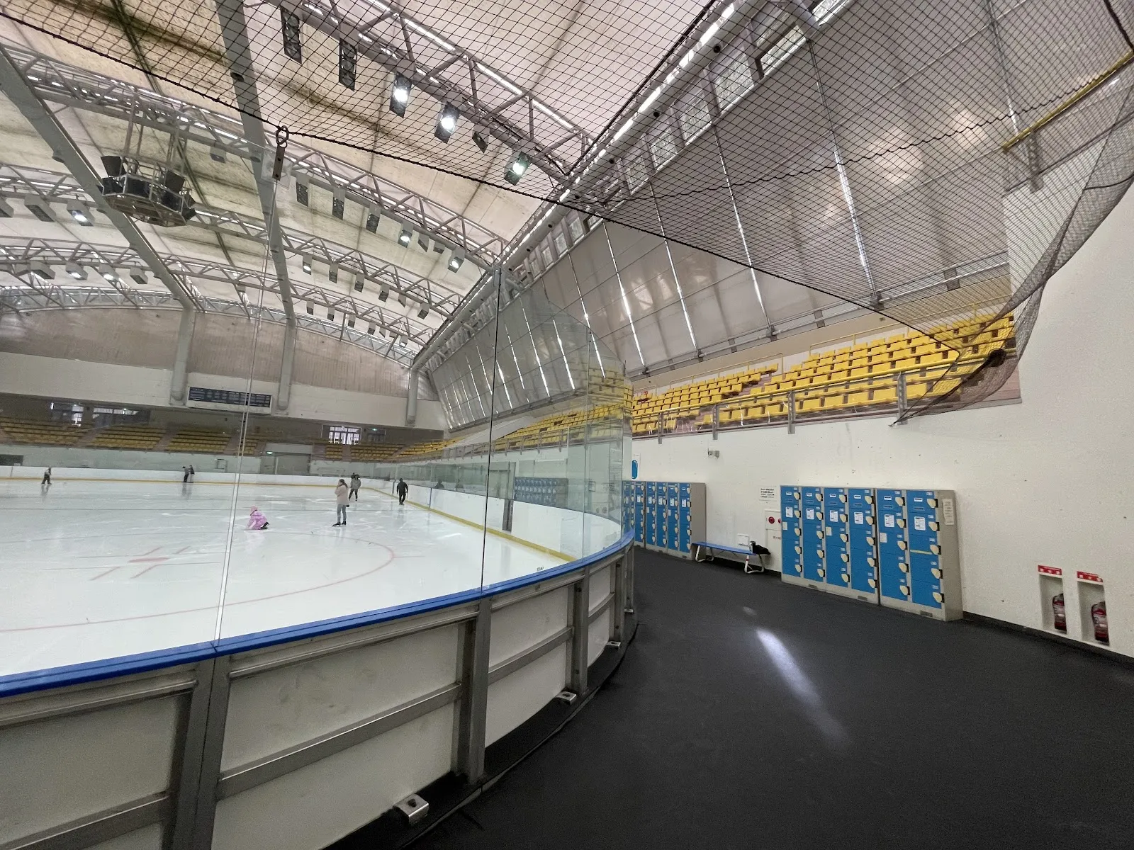 The ice sheet at Misawa Ice Arena seen along the boards, with yellow stand seating under the roof and a few skaters far off on the ice — Misawa Ice Arena