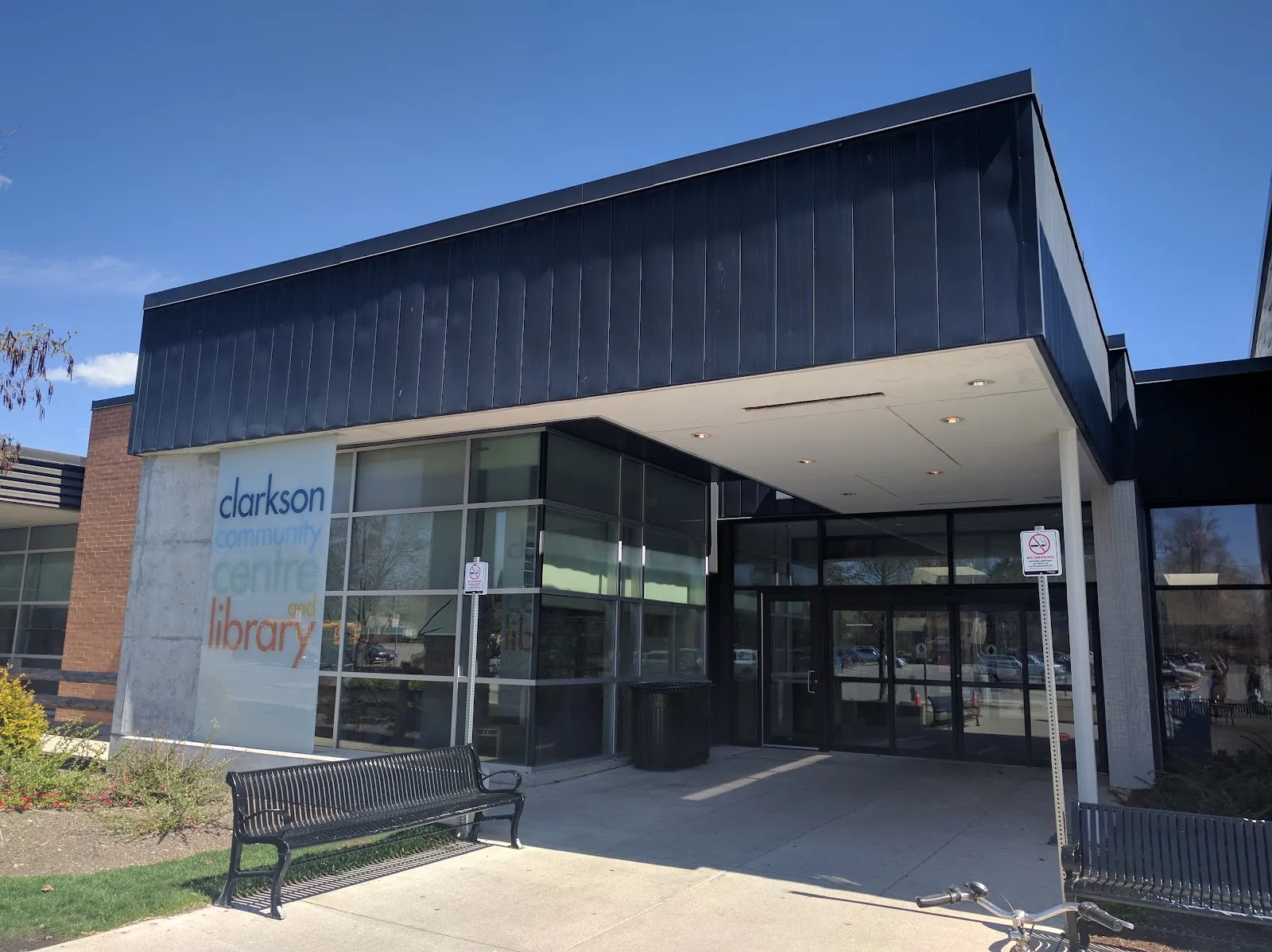 La entrada acristalada y cubierta del Clarkson Community Centre, con una marquesina de metal oscuro, un cartel de la biblioteca del centro, bancos y un aparcabicicletas, bajo un cielo azul brillante. — Clarkson Arena
