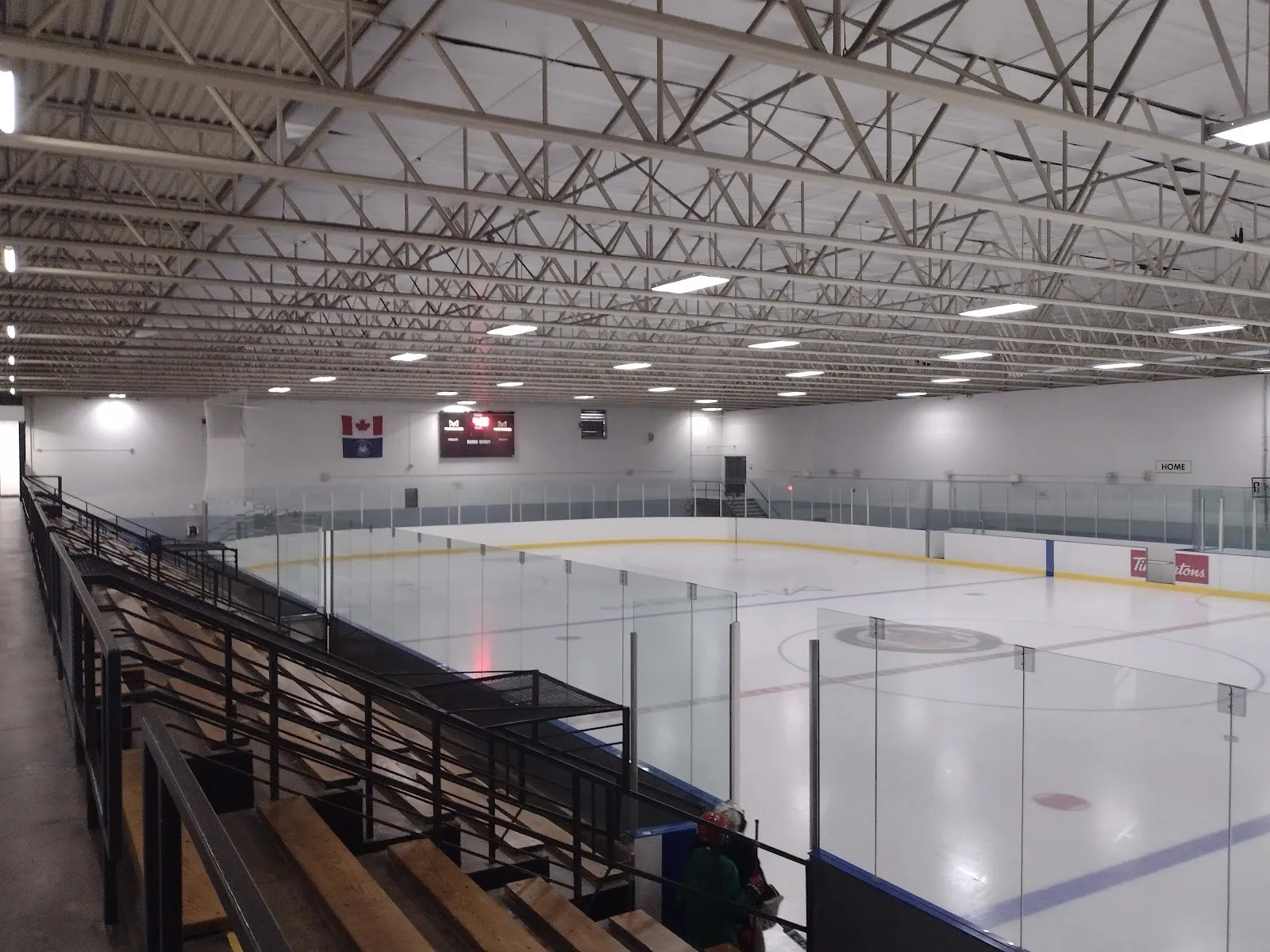 Empty indoor ice rink from the upper stands, full ice sheet with centre-ice logo, Canadian and Ontario flags and a scoreboard on the far wall, a Tim Hortons dasherboard ad, one person in gear near the bench — Clarkson Arena