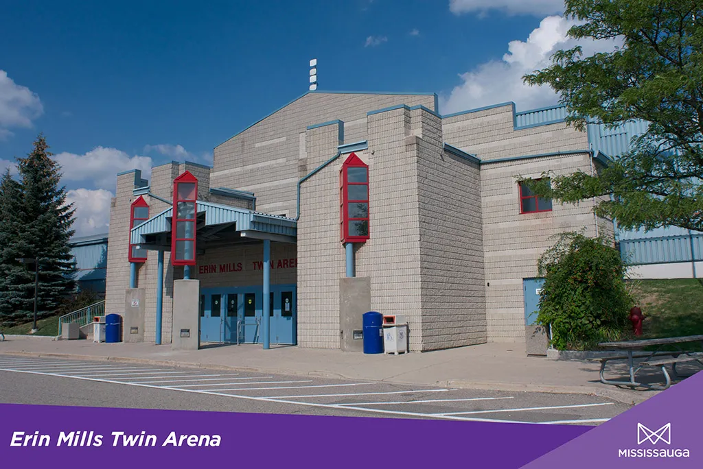 Entrada principal de Erin Mills Twin Arena, edificio de ladrillo beige con torres de tragaluz con marco rojo y detalles azules, con las letras "Erin Mills Twin Arena" sobre las puertas de vidrio, de día — Erin Mills Twin Arena