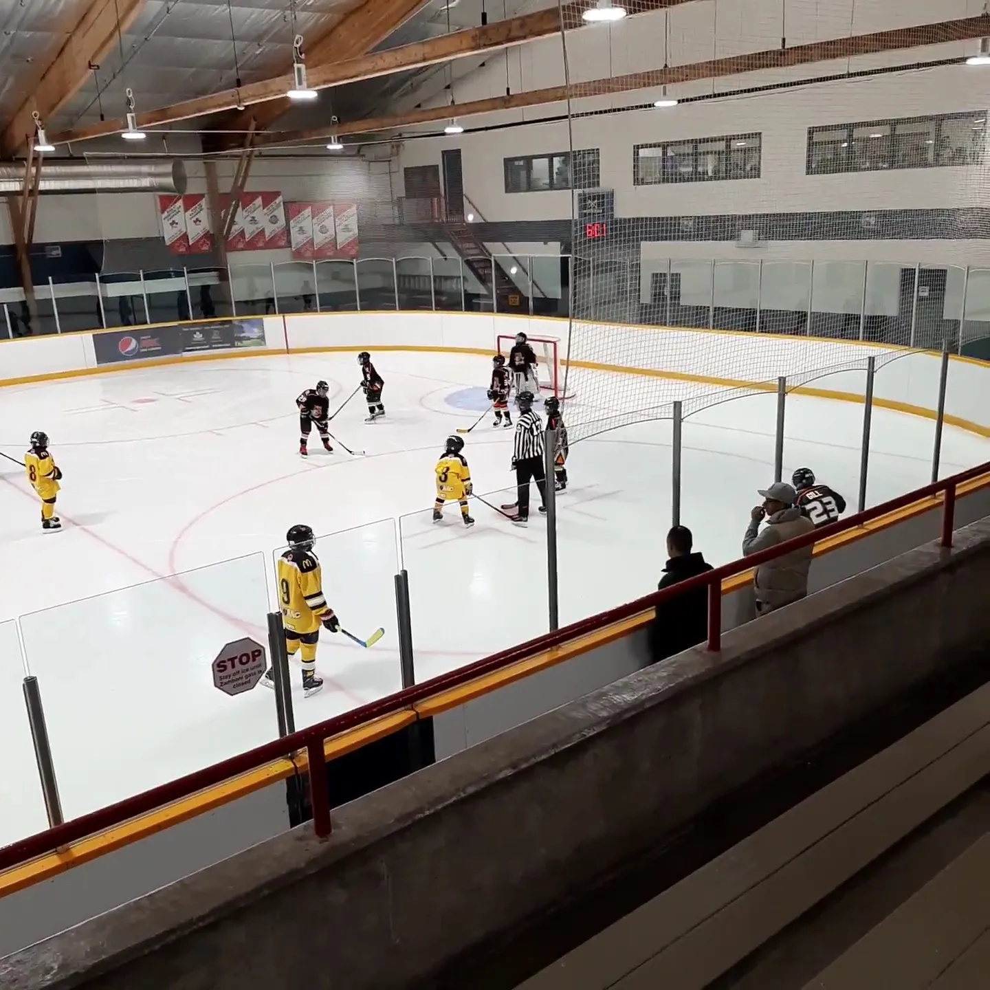 Un partido visto desde las gradas bajo un techo con vigas de madera, con jugadores repartidos por la pista y banderines en la pared del fondo. — Erin Mills Twin Arena