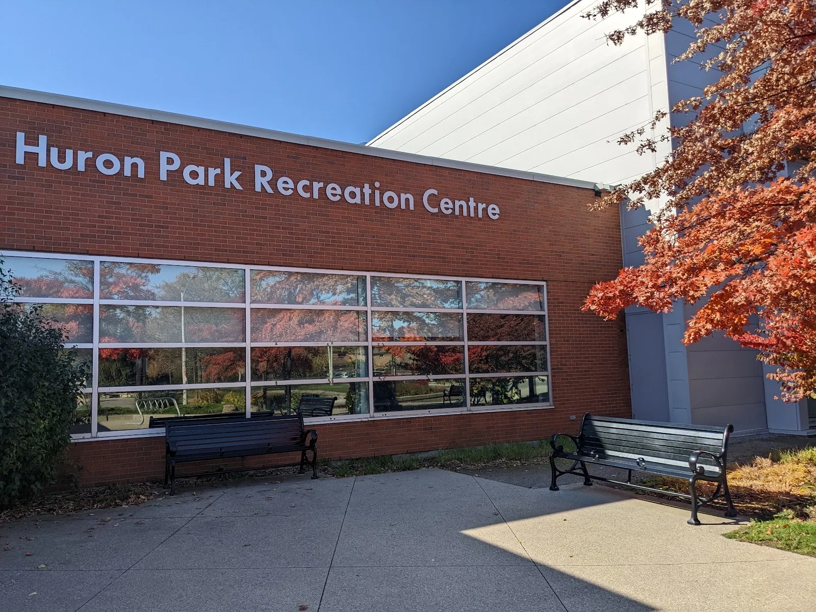 La fachada de ladrillo de Huron Park Recreation Centre con su nombre en letras blancas sobre amplias ventanas, con el alto muro de la arena detrás — Huron Park Arena