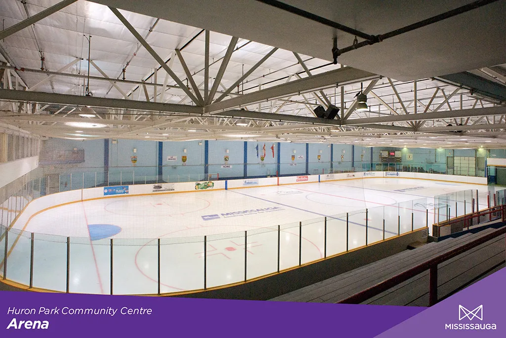 Pista de hielo vacía en Huron Park Arena, vista desde los asientos elevados para espectadores, con anuncios en las vallas y un pequeño marcador visibles al fondo. — Huron Park Arena