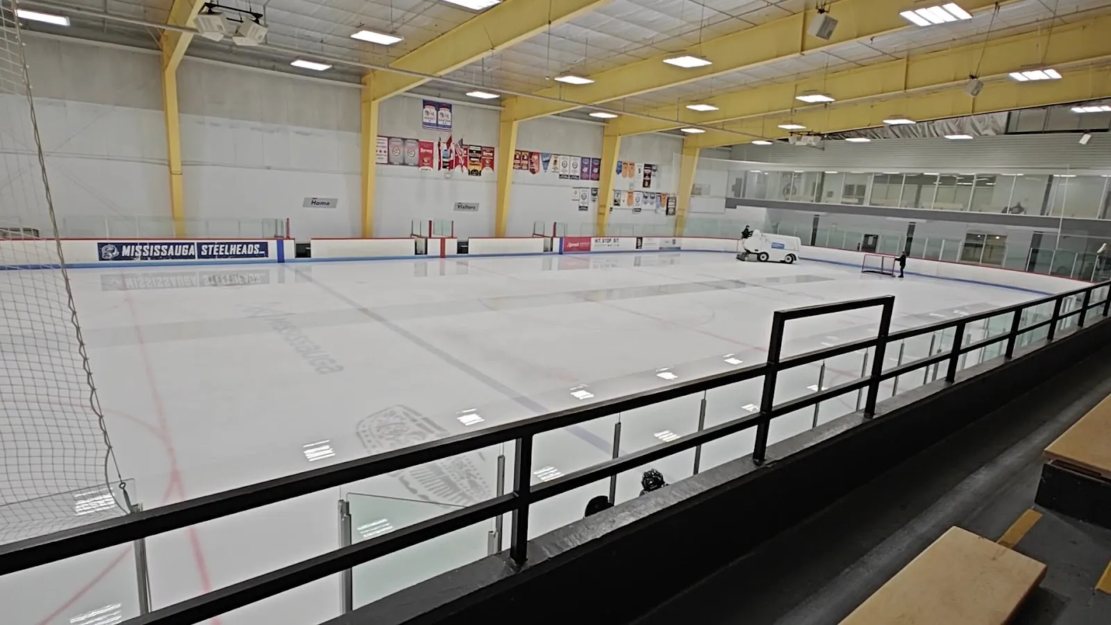 Una pista vacía de Iceland Arena desde las gradas superiores, con una pulidora de hielo cruzando el hielo recién pulido bajo vigas amarillas y mantas — Iceland Arena