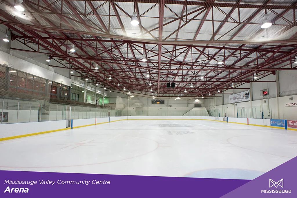 Pista de hielo vacía en la arena del Mississauga Valley Community Centre, vista desde el nivel del hielo hacia las vallas del fondo, con armaduras rojas en el techo y una manta del equipo 'Cooksville' y un letrero de 'Visitors' en las vallas. — Mississauga Valley Community Centre Arena