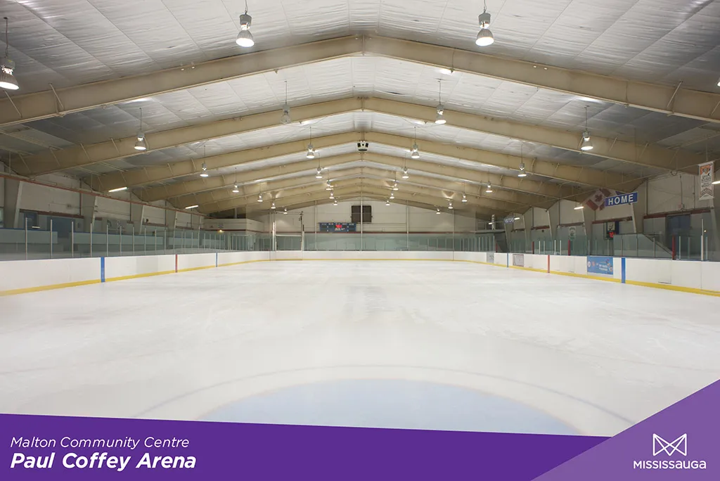 Pista de hielo vacía desde el nivel del hielo a lo largo de Paul Coffey Arena, bajo un techo a dos aguas, con un marcador y un letrero HOME en la banca. — Paul Coffey Arena