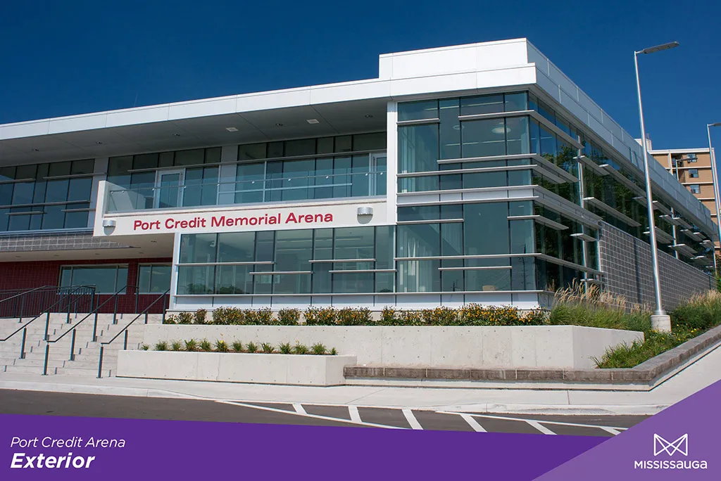 Esquina frontal de vidrio y paneles blancos de la arena en un día despejado, con un letrero rojo de "Port Credit Memorial Arena" en la pared sobre la entrada principal y un acceso escalonado de concreto. — Port Credit Memorial Arena
