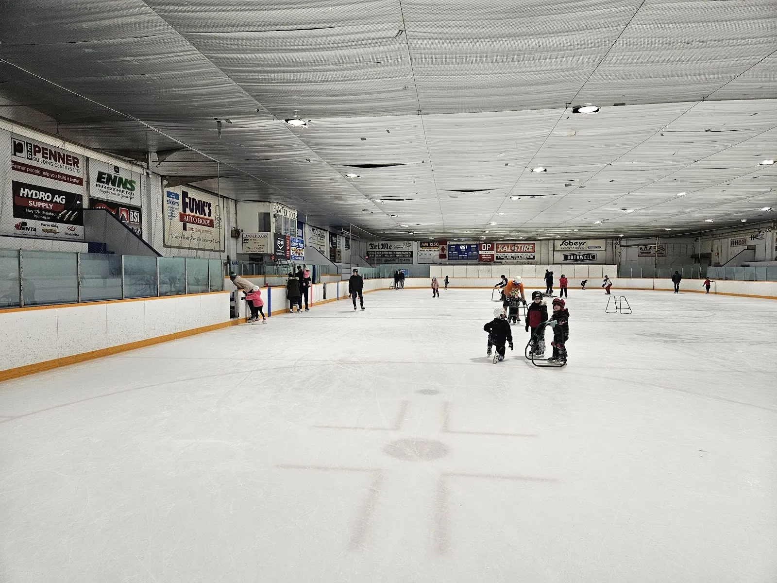 Una pista de hielo amplia con unos cuantos patinadores bajo un techo blanco aislado, con anuncios de patrocinadores y una cabina de prensa en el lado opuesto. — Mitchell Arena