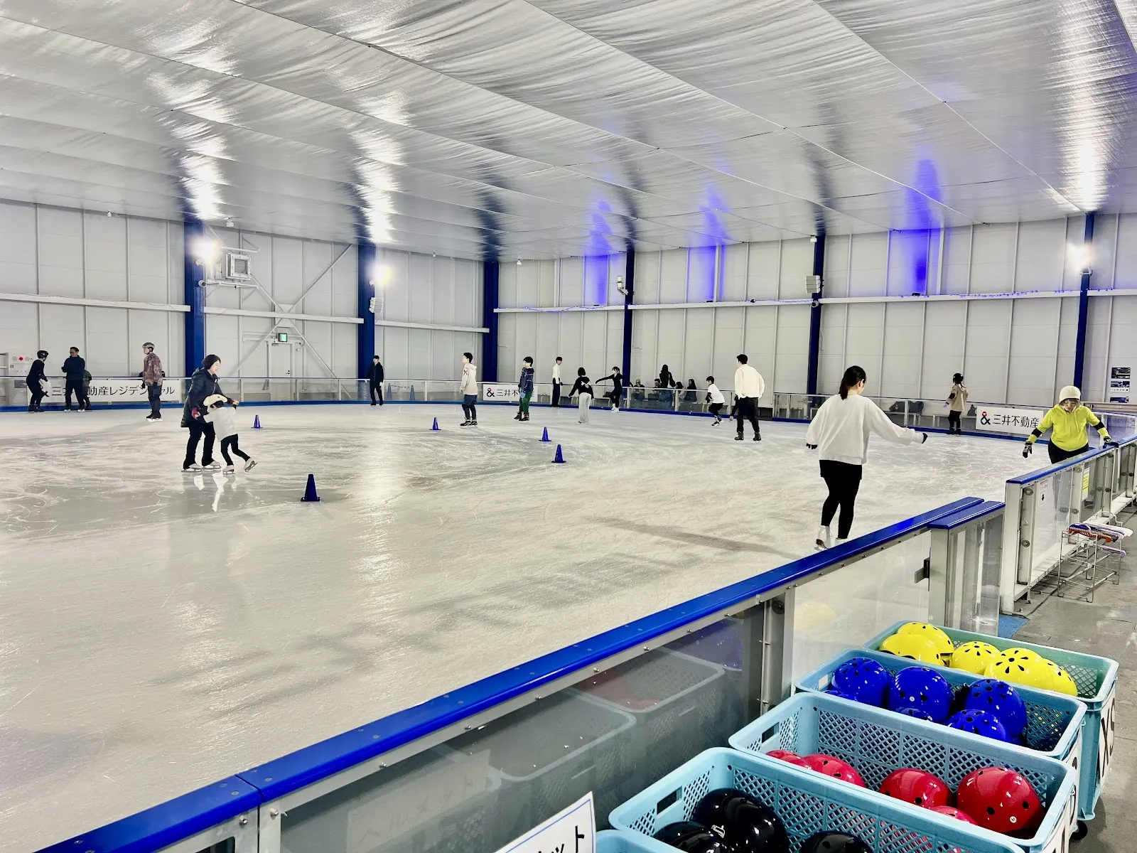 フェンス際から見た、三井不動産アイスパーク船橋の氷面。右下にヘルメットのかごがあり、青い柱と、遠くに数人の滑走者が見えます — Mitsui Fudosan Ice Park Funabashi
