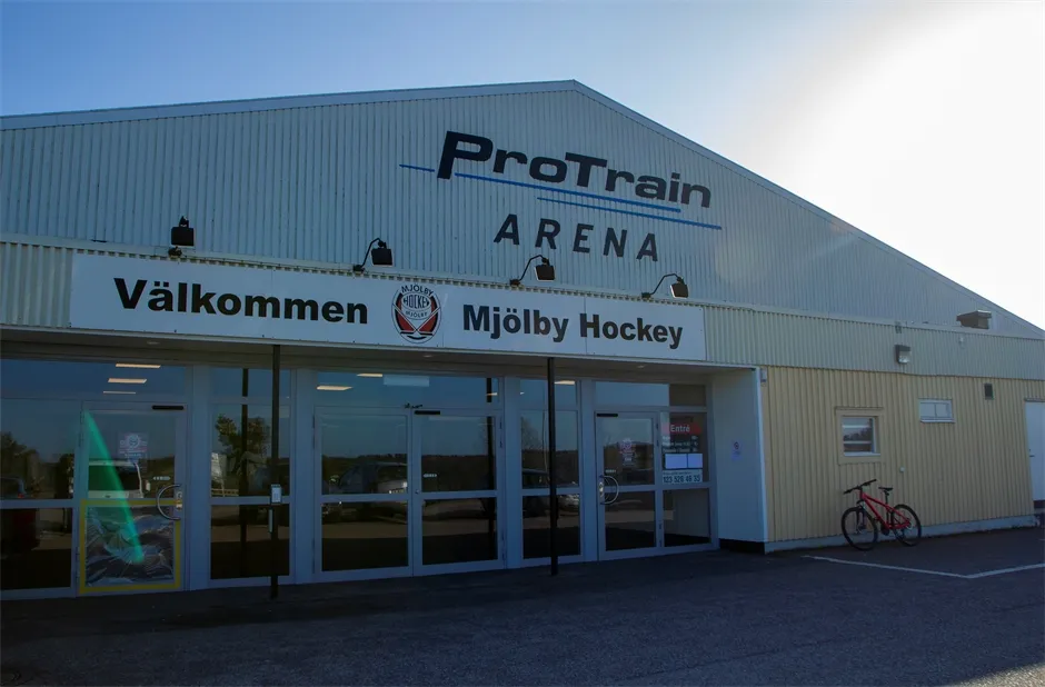 The front of the ProTrain Arena in Mjölby with the arena name on the gable, the Mjölby Hockey welcome sign over the glass entrance and a bicycle at the wall — Mjölby ishall (ProTrain Arena)
