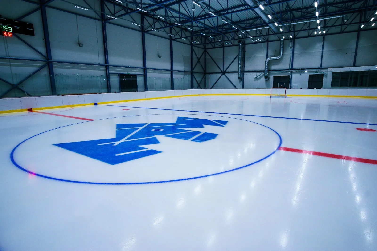 Empty ice sheet in the MM Aréna hall with the blue MM logo at centre ice, the scoreboard and the steel roof. — MM Aréna