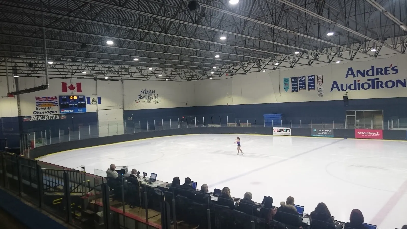 A wide view across the sheet from the stands with a lone skater and spectators at the near rail under a white roof lined with banners. — MNP Place