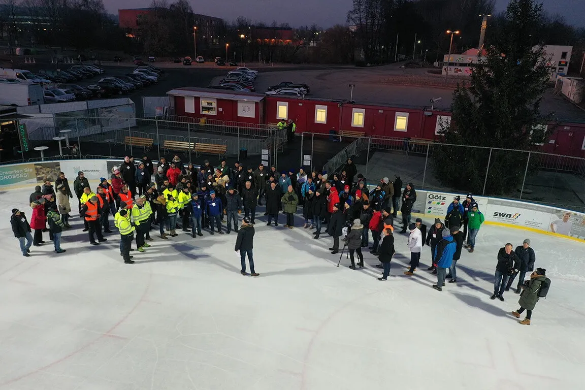 The mobile ice rink at dusk seen from above, a large group of people standing on the white ice beside red container huts, with parked vehicles and trees behind. — Mobile Kunsteislaufanlage Neumarkt
