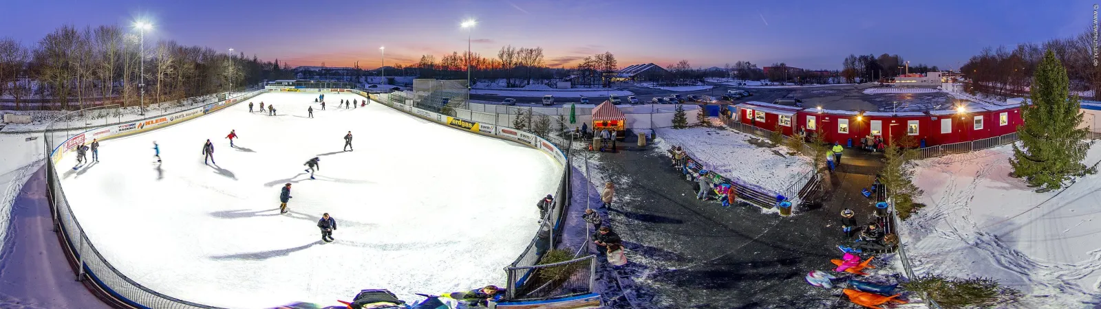 A wide dusk view of the floodlit mobile ice rink on the Volksfestplatz, with skaters across the ice, the red cabin row and the Christmas tree beside it. — Mobile Kunsteislaufanlage Neumarkt
