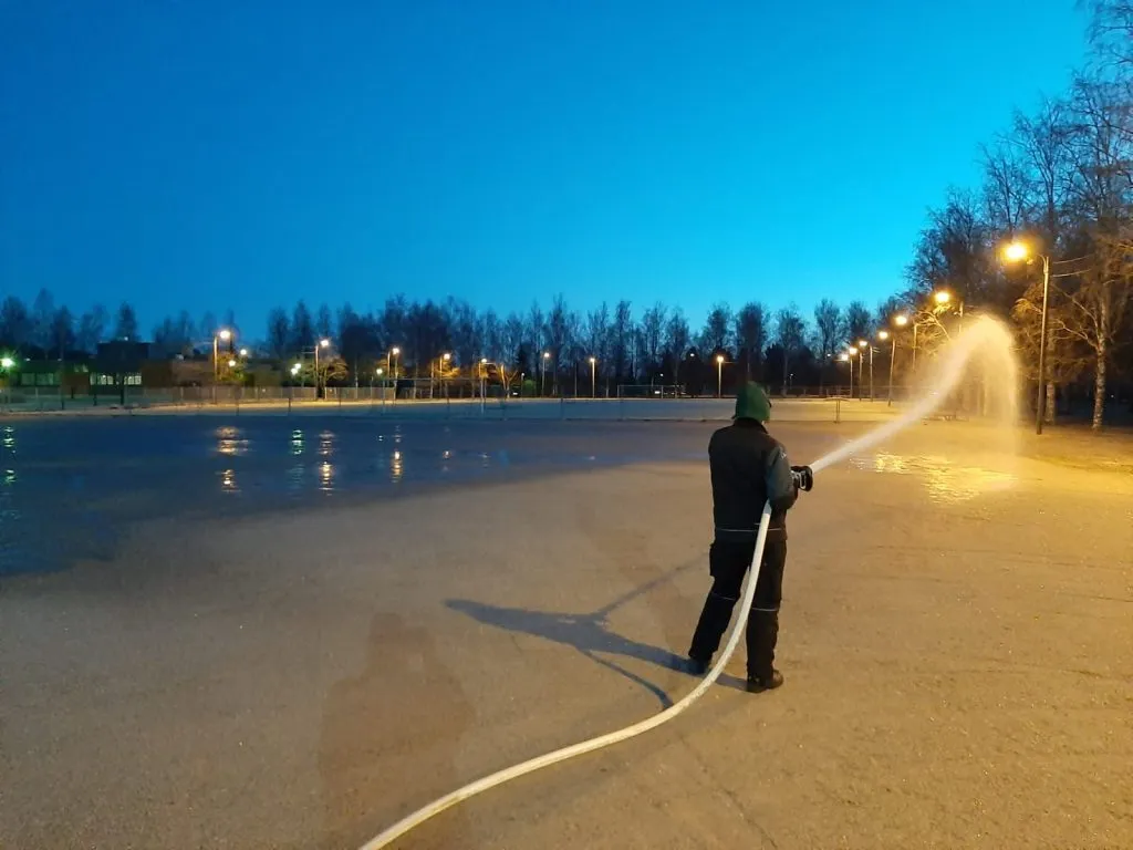 En arbetare som översvämmar rinken vid Moisio med en slang i skymningen, sedd bakifrån, med gatlyktorna tända runt planen. – Moision kaukalo ja luistelukenttä (Salo)